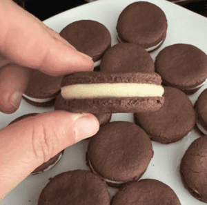 Caucasian fingers holding chocolate Oreo cookies sandwich filled with white buttercream icing over a tray of other filled Oreo sandwich cookies that are resting on a white plate.