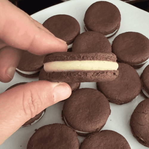 Caucasian fingers holding chocolate Oreo cookies sandwich filled with white buttercream icing over a tray of other filled Oreo sandwich cookies that are resting on a white plate.