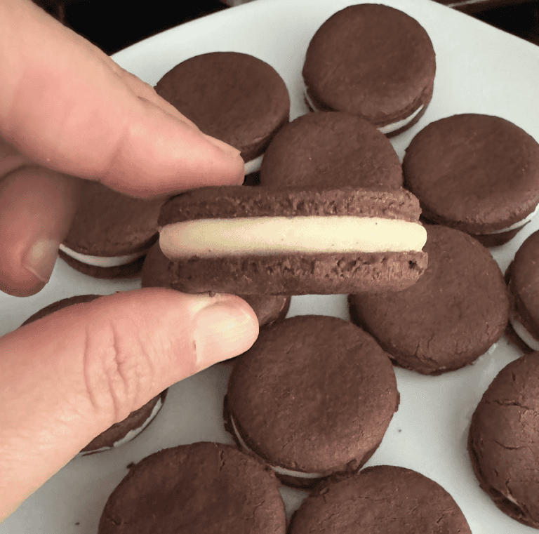Caucasian fingers holding chocolate Oreo cookies sandwich filled with white buttercream icing over a tray of other filled Oreo sandwich cookies that are resting on a white plate.