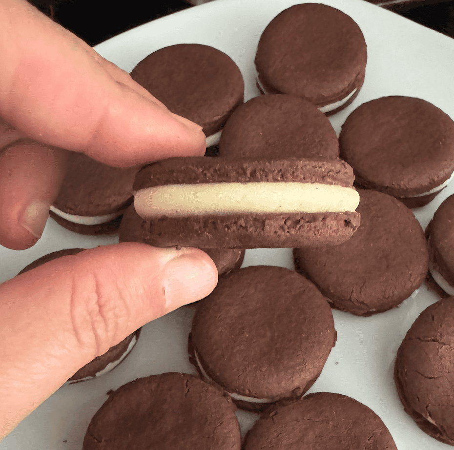 A hand holding a small brown cookie with while filling in the middle and several more cookies in the background on a white plate