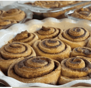 Trays of fresh milled gluten free cinnamon rolls. 9 are in a white ceramic pie dish lined with brown coming. There is another 9 (3 visible) are in a blue ceramic pie dish lined with brown coming. Two more glass dishes are in the background filled with cooked cinnamon rolls. They are brown with dark brown maple syrup/butter swirls.