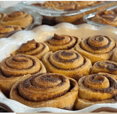 Trays of fresh milled gluten free cinnamon rolls. 9 are in a white ceramic pie dish lined with brown coming. There is another 9 (3 visible) are in a blue ceramic pie dish lined with brown coming. Two more glass dishes are in the background filled with cooked cinnamon rolls. They are brown with dark brown maple syrup/butter swirls.