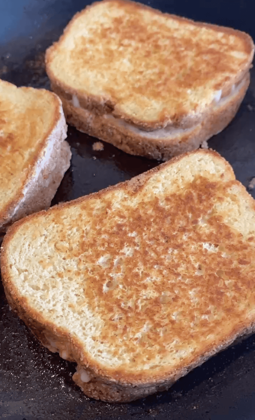 Three golden-brown grilled cheese sandwiches on gluten free sandwich bread in a black cast iron pan.