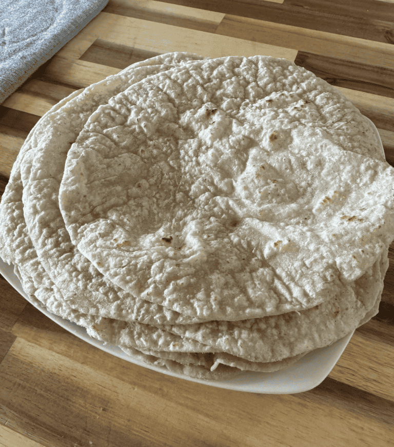 6 tan/speckled with dark brown cooked areas - gluten free tortilla wraps on a white plate sitting on an acacia wood countertop. There is a grey drying mat on the left side in the background.