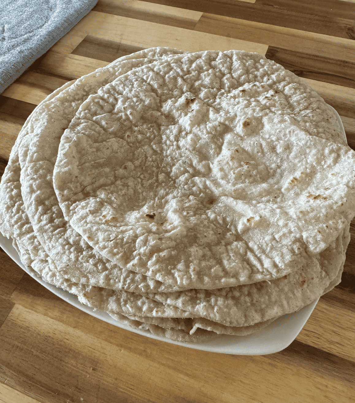 6 tan/speckled with dark brown cooked areas - gluten free tortilla wraps on a white plate sitting on an acacia wood countertop. There is a grey drying mat on the left side in the background.