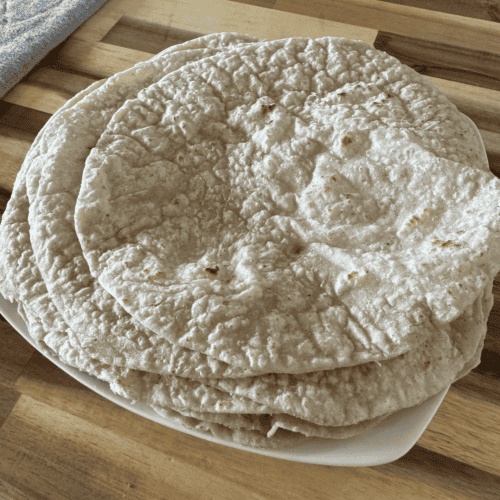 6 tan/speckled with dark brown cooked areas - gluten free tortilla wraps on a white plate sitting on an acacia wood countertop. There is a grey drying mat on the left side in the background.