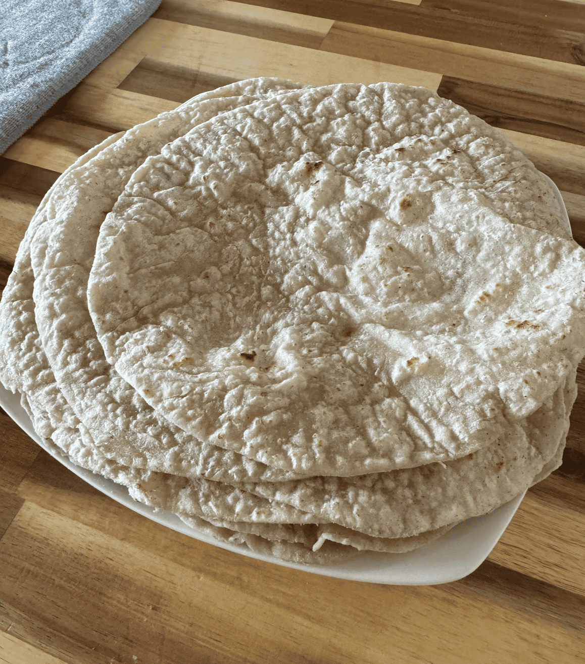 6 tan/speckled with dark brown cooked areas - gluten free tortilla wraps on a white plate sitting on an acacia wood countertop. There is a grey drying mat on the left side in the background.