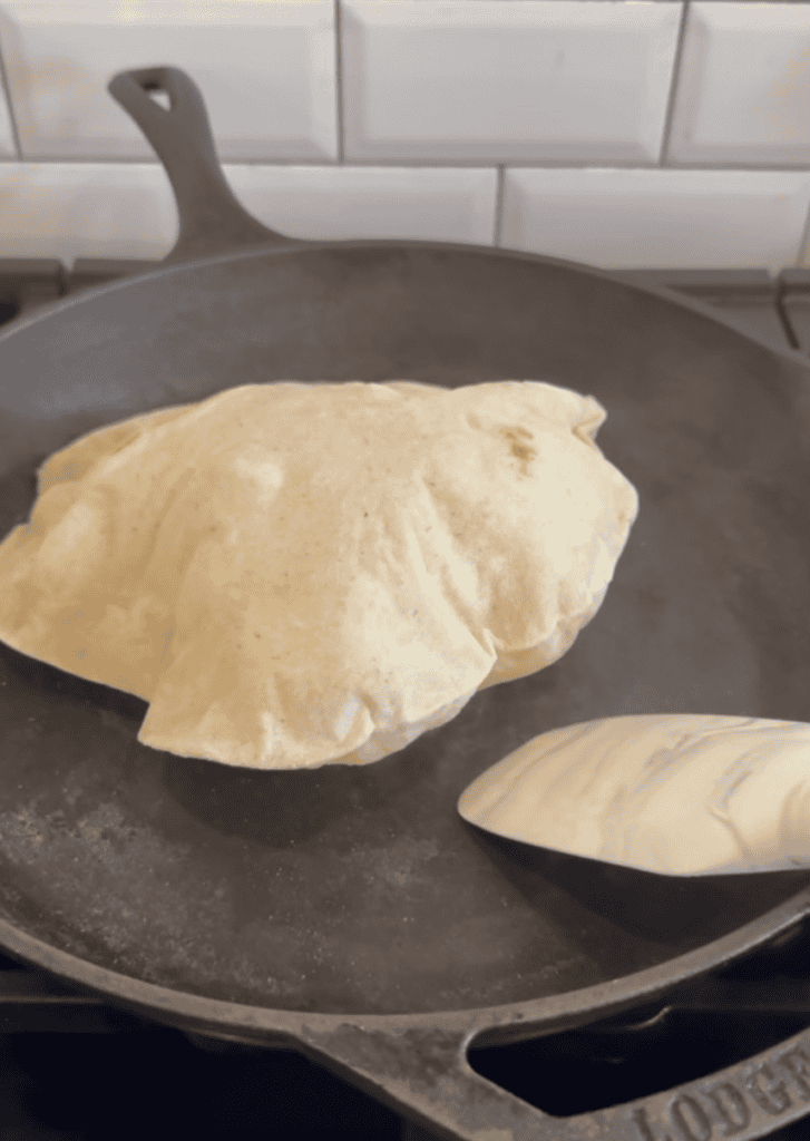 A gluten free tortilla bubbling up due to high heat in a black cast iron skillet sitting on a black stove grate. White Subway tile is in the background