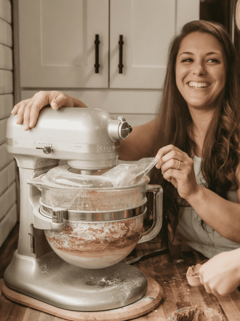 Cookbook author, Jennifer Roberts, is mixing chocolate icing. Her son is holding a scoop of cocoa powder over a messy counter. The mixer is a silver kitchen aid professional with a glass bowl. Jennifer is smiling showing her teeth at her son as she holds onto the machine while it mixes. She is wearing light blue overalls with a white t-shirt underneath. There is a knife on the countertop. The countertop is wood grains (acacia). The tile on the wall is subway tile and Jennifer has a white cabinet with black handles behind her.