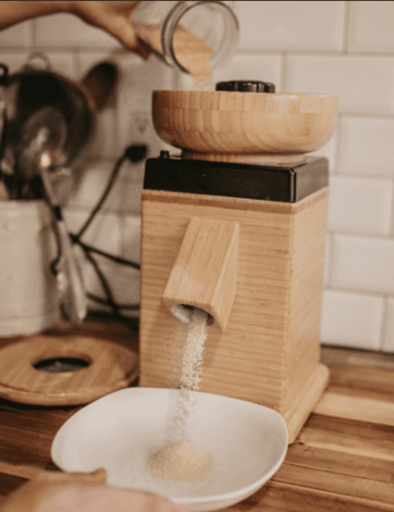 Using the harvest grain mill from nutrimill while milling Millet flour. using a glass mason jar to pour the millet flour. The lid of the grain mill is on a wooden counter along with the white bowl that is catching the fresh milled flour.