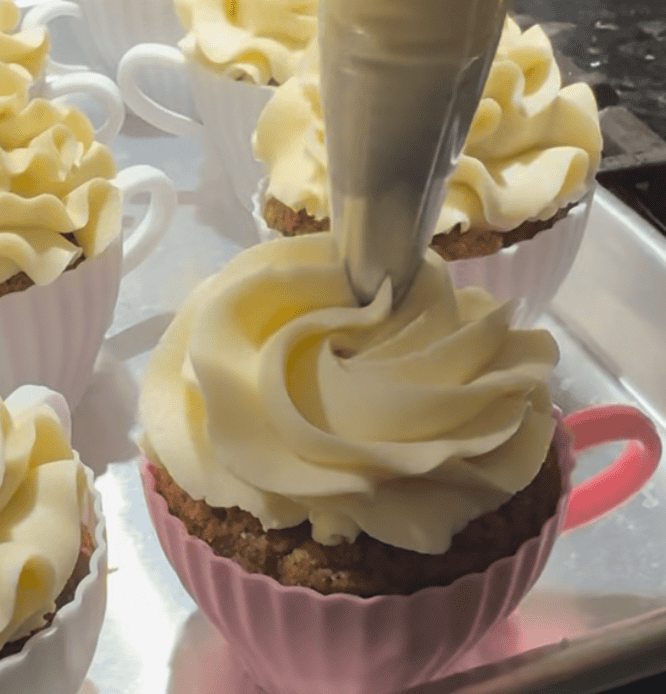 A silicone cupcake mold shaped like a tea cup with cupcakes baked into them placed on a baking metal cooking sheet. The cupcake is being piped with buttercream icing.