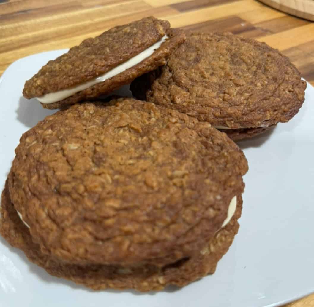 A white plate with 3 large round brown cookies with white filling inside on a wood counter top 