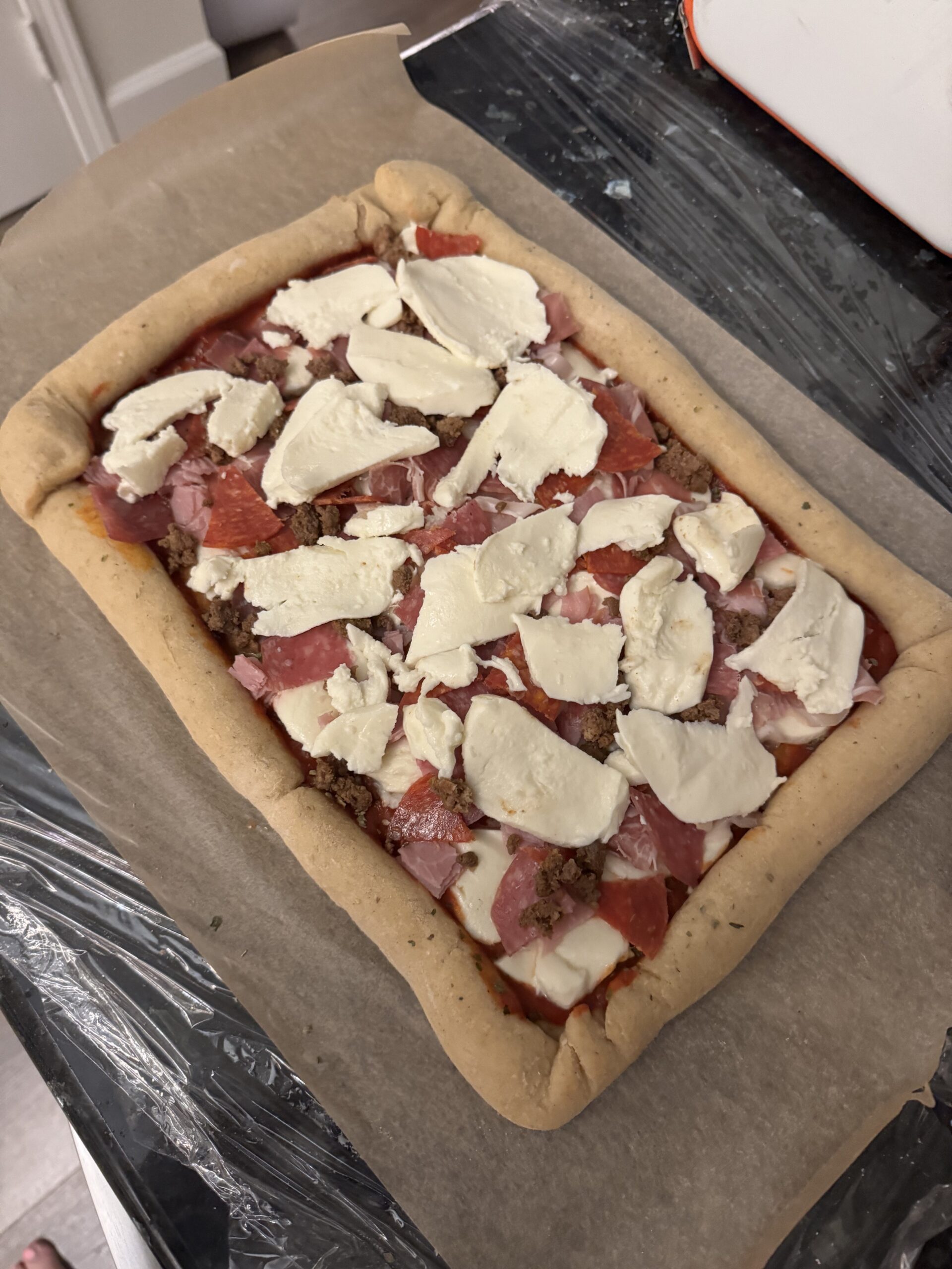 A square of brown parchment paper with a square pizza dough with red sauce, slices of mozzarella, bacon and pepperoni shreds and ground meat