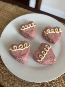 Four pink heart cakes on a white plate that is place on top of a burlap table runner. The pink hearts have three to four white squiggle loops on them for decoration.
