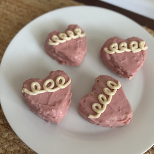 Four pink heart cakes on a white plate that is place on top of a burlap table runner. The pink hearts have three to four white squiggle loops on them for decoration.