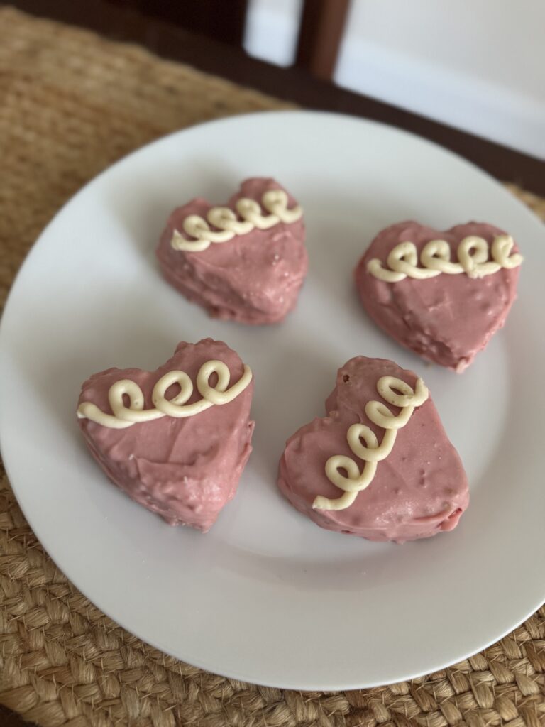 Four pink heart cakes on a white plate that is place on top of a burlap table runner. The pink hearts have three to four white squiggle loops on them for decoration.