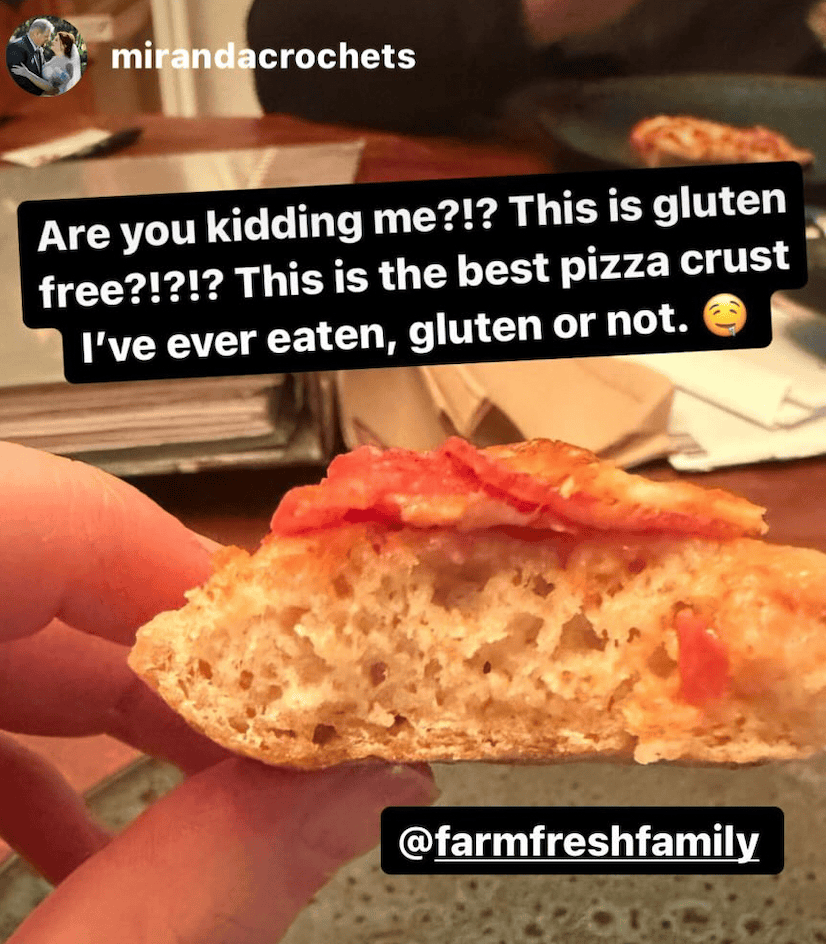 A hand holding a piece of pizza dough with cheese and pepperoni and black text bubble with white text reading Are you kidding me?!? This is gluten free?!?!?! This is the best pizza crust I've ever eaten, gluten or not. crying laughing emoji and @farmfreshfamily below the pizza 