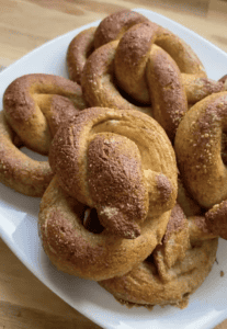 A pile of 6 soft, fluffy gluten free pretzels on a white plate sitting on a wooden countertop.