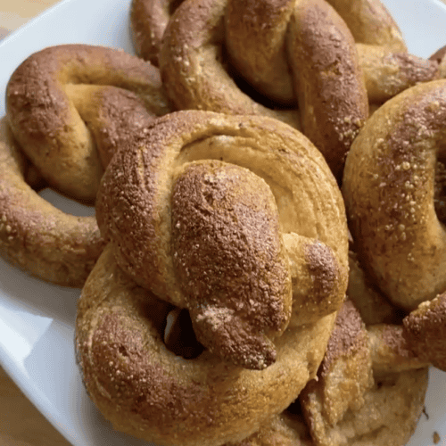 A pile of 6 soft, fluffy gluten free pretzels on a white plate sitting on a wooden countertop.