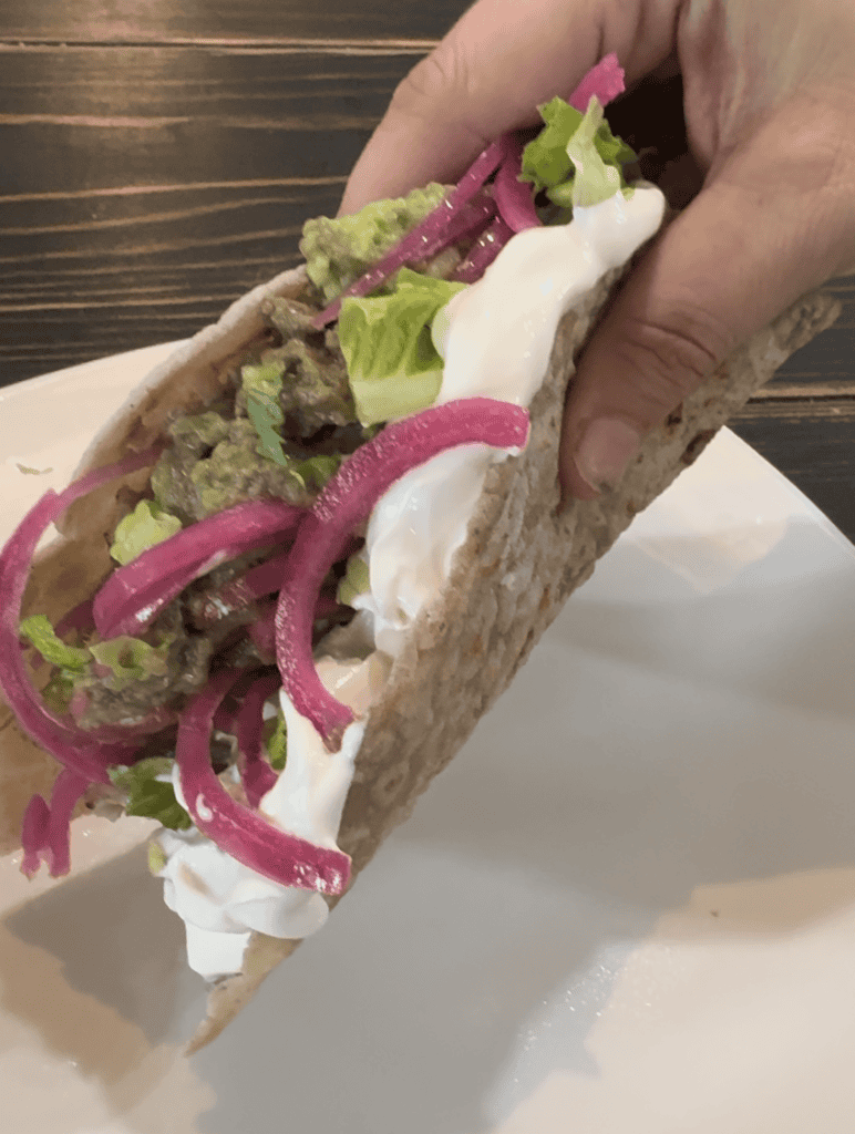 Hand holding a soft shell taco with beef, guacamole, pickled onions, sour cream and shredded lettuce
