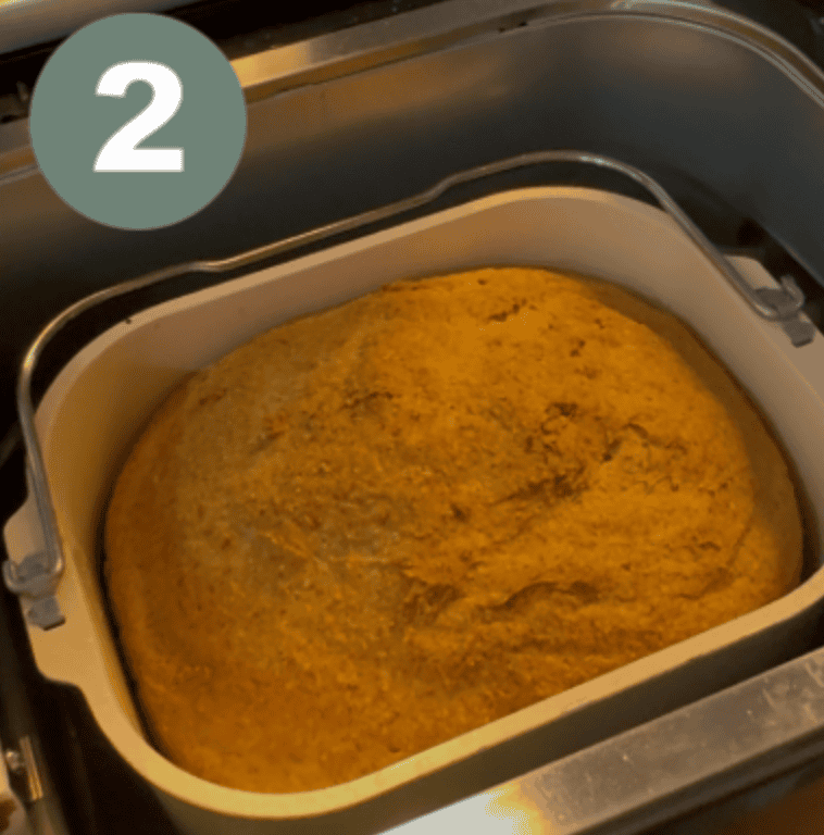 A risen ball of dough in a square bread maker pan with a green circle and a white number two in the left upper corner 