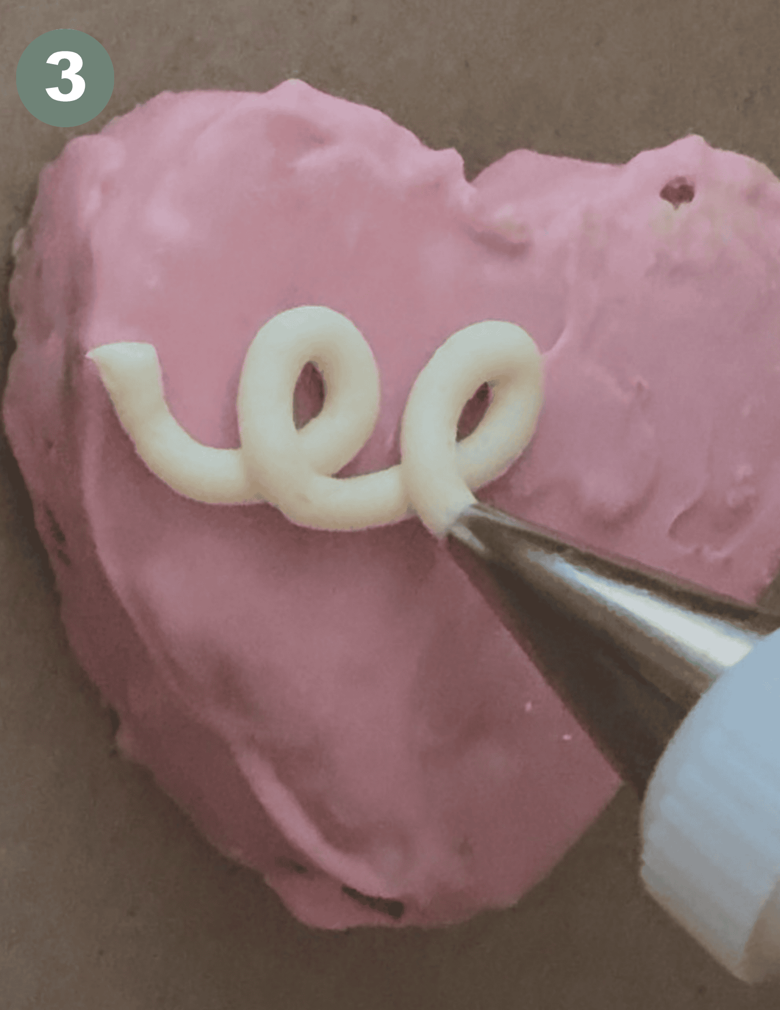 A pink frosted heart with a metal tip of frosting and two white swirls in the middle of the heart