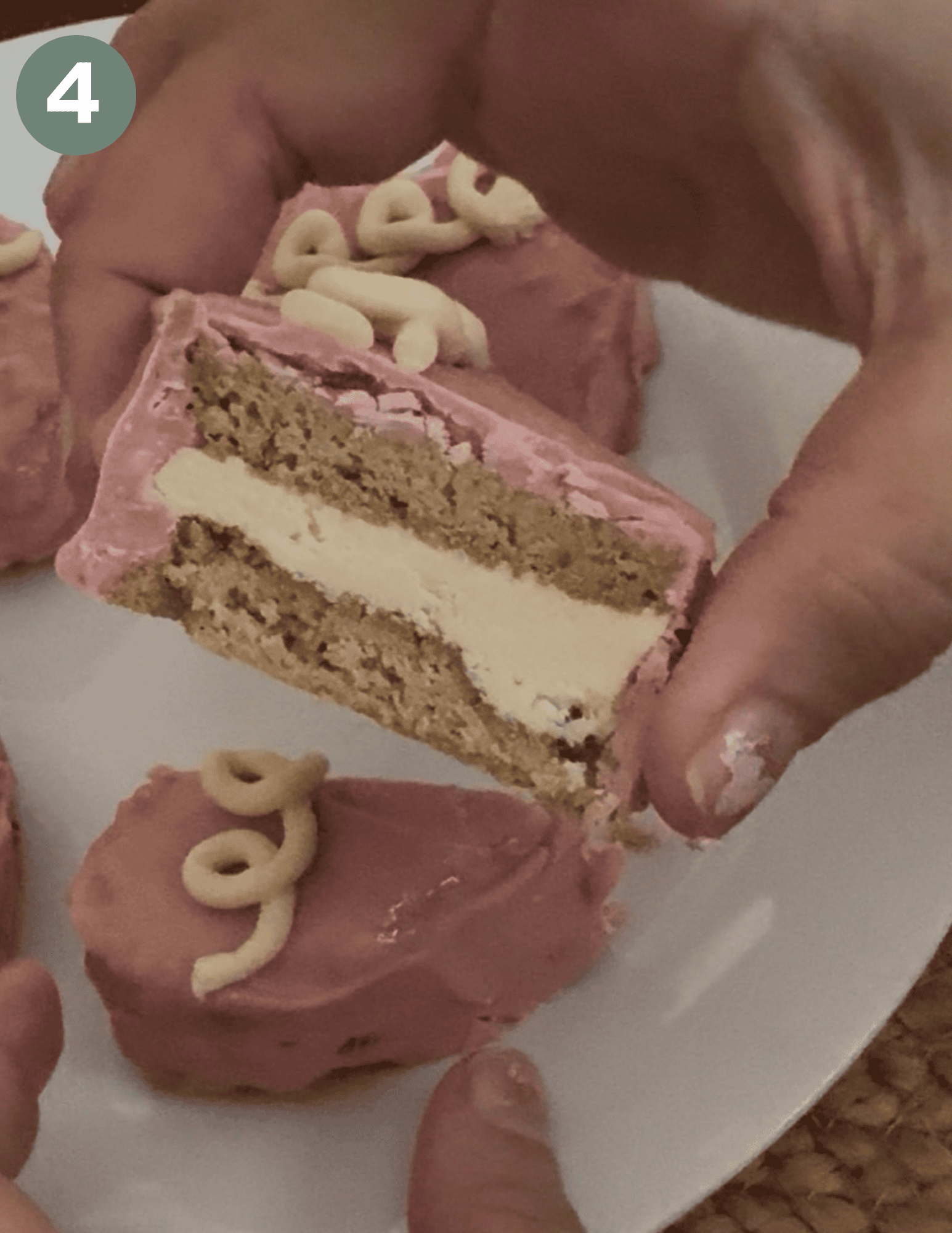 A hand holding a small cake with pink frosting on the outside, two layers of brown yellow cake and white frosting in the middle. In the background a half of a pink heart cake with swirls and a green circle with a white number 4 in the upper left hand corner