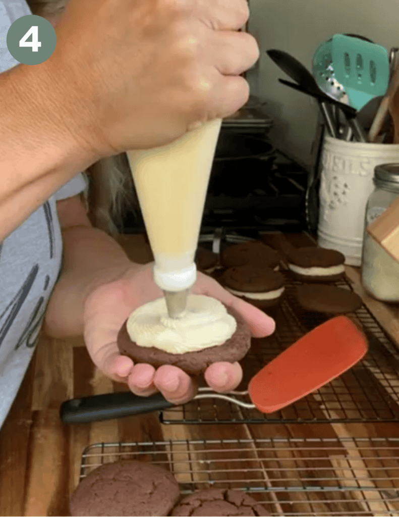 A hand holding a brown cookie and another hand applying white icing to the cookie with more cookies and kitchen utensils in the background and a small green circle with a white number four in the upper left hand corner