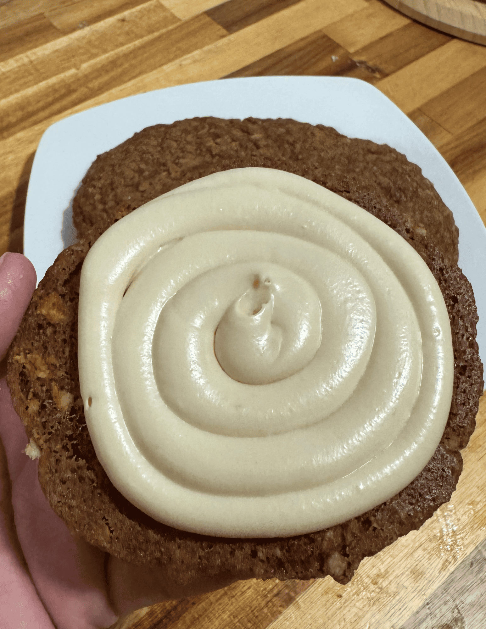 A hand holding a large round brown cookie with white icing covering the cookie in front of more cookies on a plate on a wooden countertop