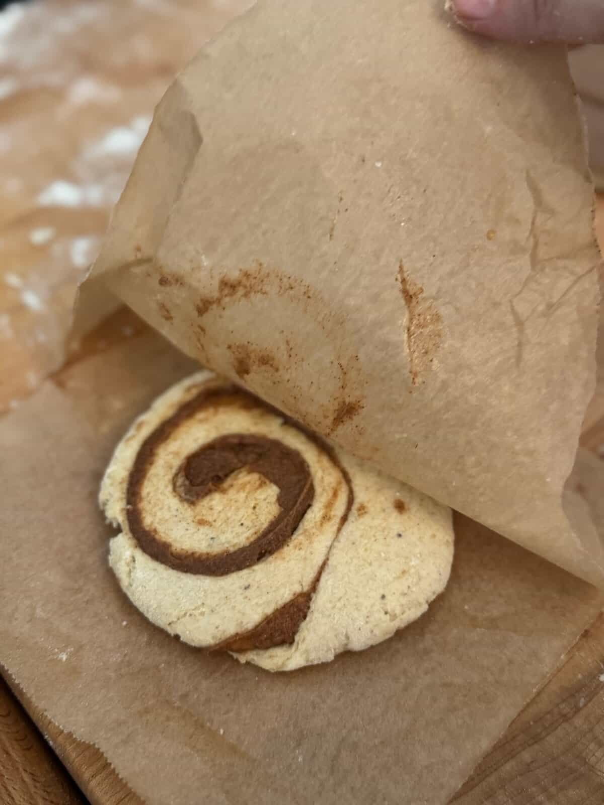 A round piece of raw gluten free honey bun dough on a piece of parchment paper with a hand pulling back another piece of parchment paper from the top