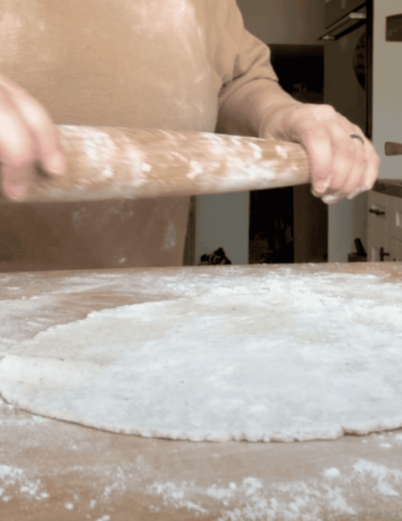 A person rolling dough very thinly with a wooden rolling pin on a wooden counter top